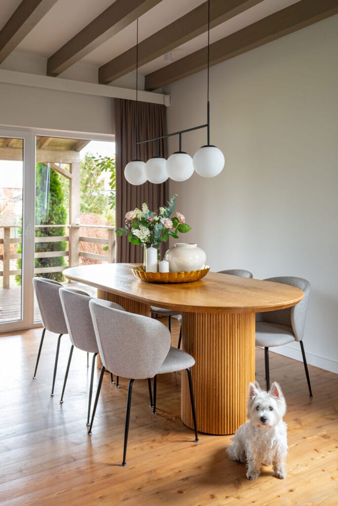 Elegancka jadalnia z dużym, drewnianym stołem otoczonym pięcioma tapicerowanymi krzesłami. Nad stołem zawieszona jest nowoczesna lampa z kulistymi kloszami. W tle drewniane belki sufitowe i zasłony w ciepłych odcieniach.