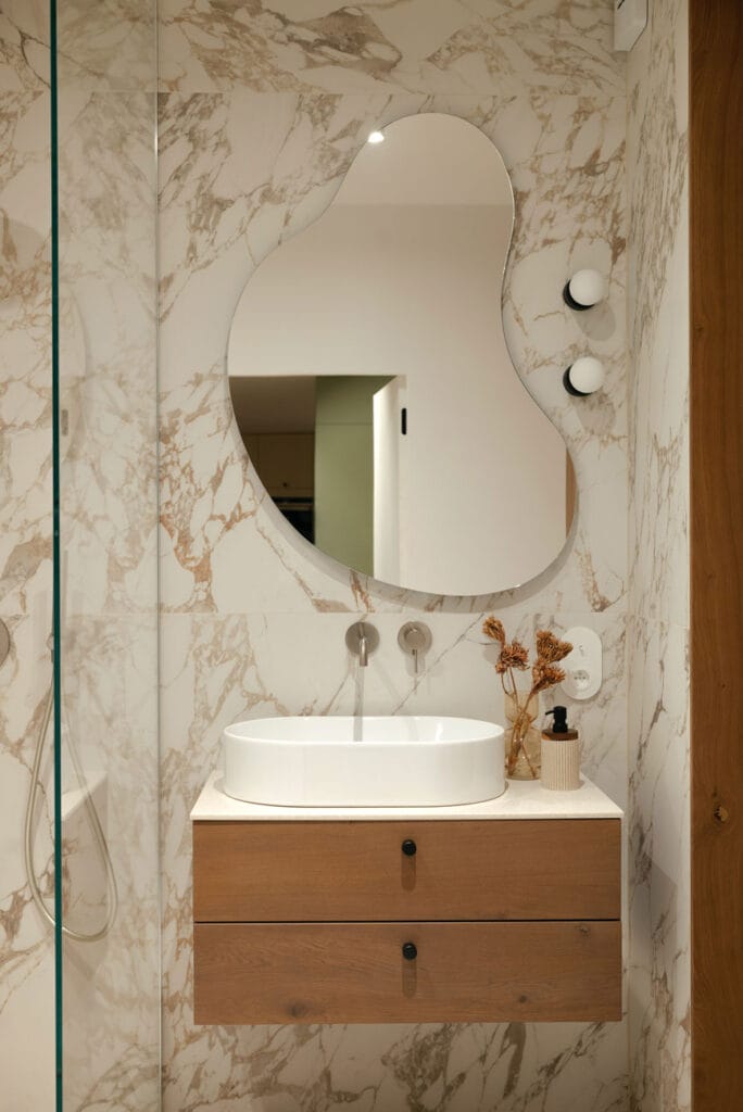 Elegancka łazienka z marmurowymi ścianami, drewnianą szafką pod umywalką i designerskim lustrem o nieregularnym kształcie.