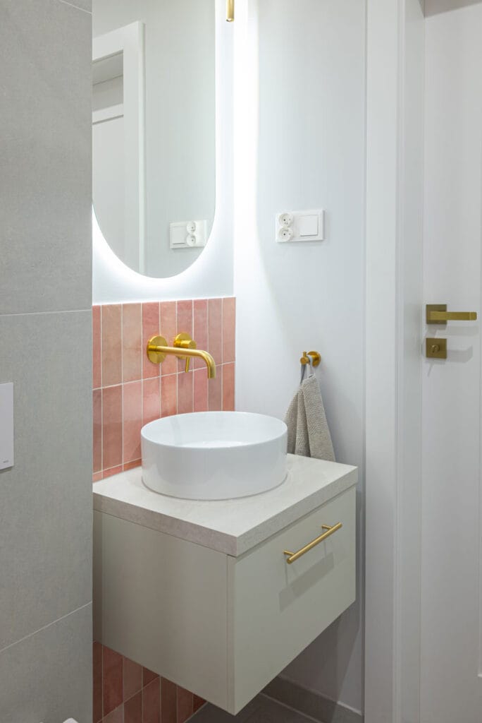 Stylowa łazienka z podświetlanym okrągłym lustrem, złotą armaturą i pastelowymi różowymi płytkami nad umywalką.