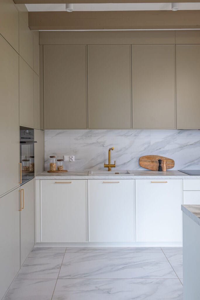Nowoczesna kuchnia w jasnych barwach z wysokimi szafkami w ciepłym beżu i białymi frontami dolnych szafek. Blat i ściana wykończone są imitacją marmuru, a złote uchwyty i bateria kuchenna dodają elegancji.