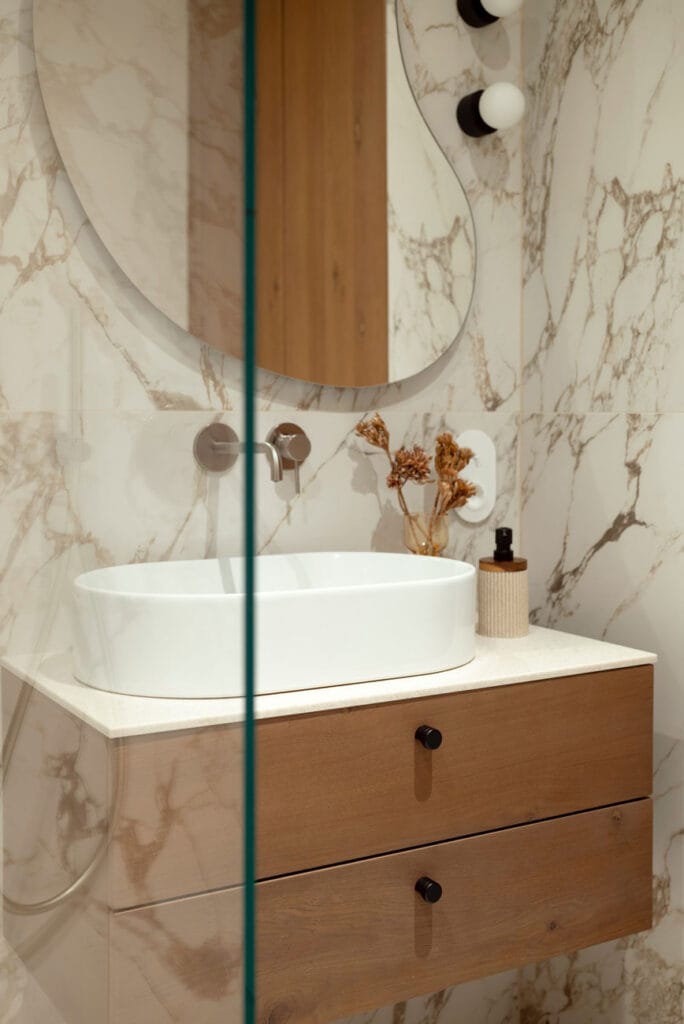 Luksusowa łazienka z dużym designerskim lustrem, marmurowymi ścianami i podwieszaną drewnianą szafką z umywalką nablatową.