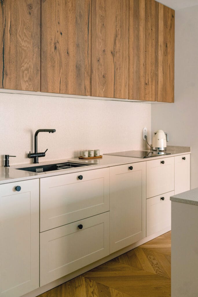 Minimalistyczna kuchnia z drewnianymi szafkami, jasnym blatem i czarnymi uchwytami, utrzymana w eleganckim stylu.