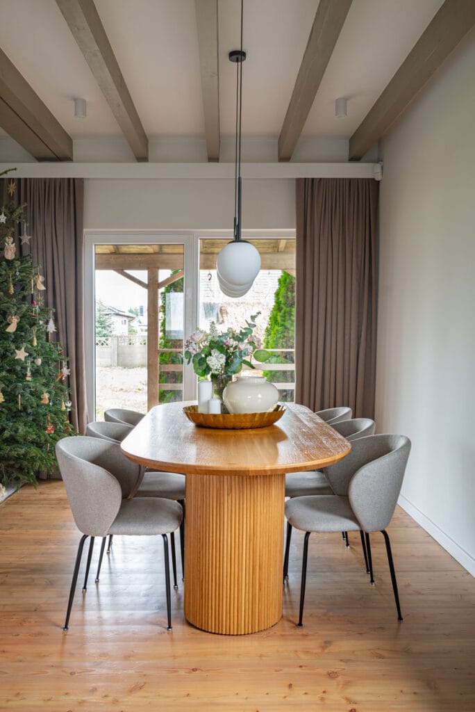 Jadalnia z dużym, drewnianym stołem o zaokrąglonych krawędziach i szarymi, tapicerowanymi krzesłami. W tle choinka, duże okna z widokiem na ogród i drewniane belki na suficie.