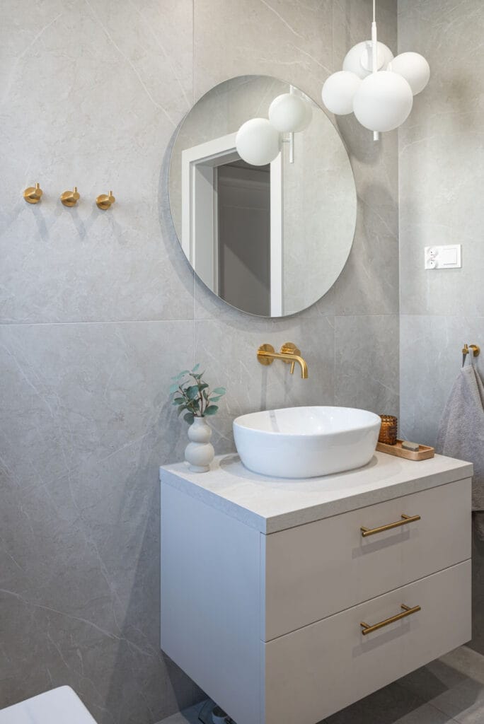 Nowoczesna łazienka z dużym, okrągłym lustrem, złotą armaturą i wiszącą lampą w kształcie kul.