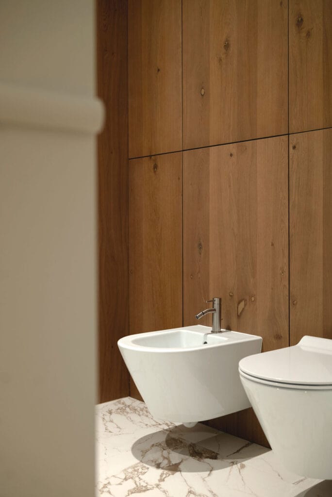 Nowoczesna toaleta z bidetem i WC na tle ściany wykończonej naturalnym drewnem, na marmurowej podłodze.