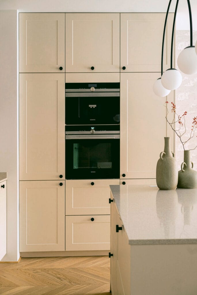 Nowoczesna zabudowa kuchenna w odcieniach beżu z czarnymi detalami i podwójnym piekarnikiem.