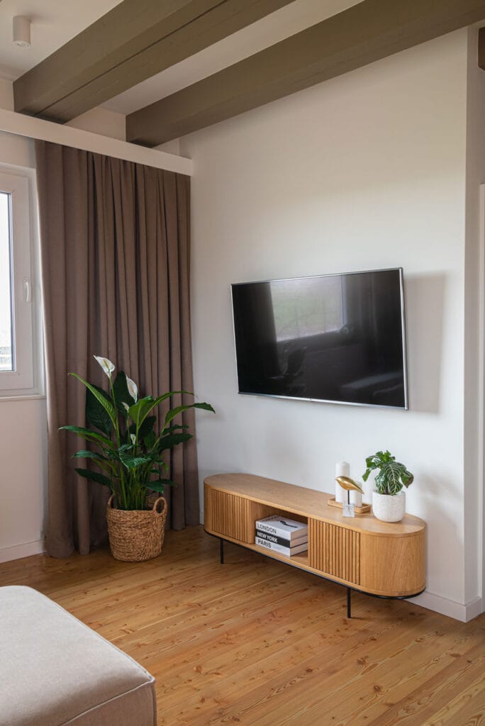 Nowoczesny salon z telewizorem zawieszonym na ścianie, elegancką drewnianą komodą na czarnych nóżkach i rośliną w plecionym koszu. W tle długie, beżowe zasłony i jasna podłoga.
