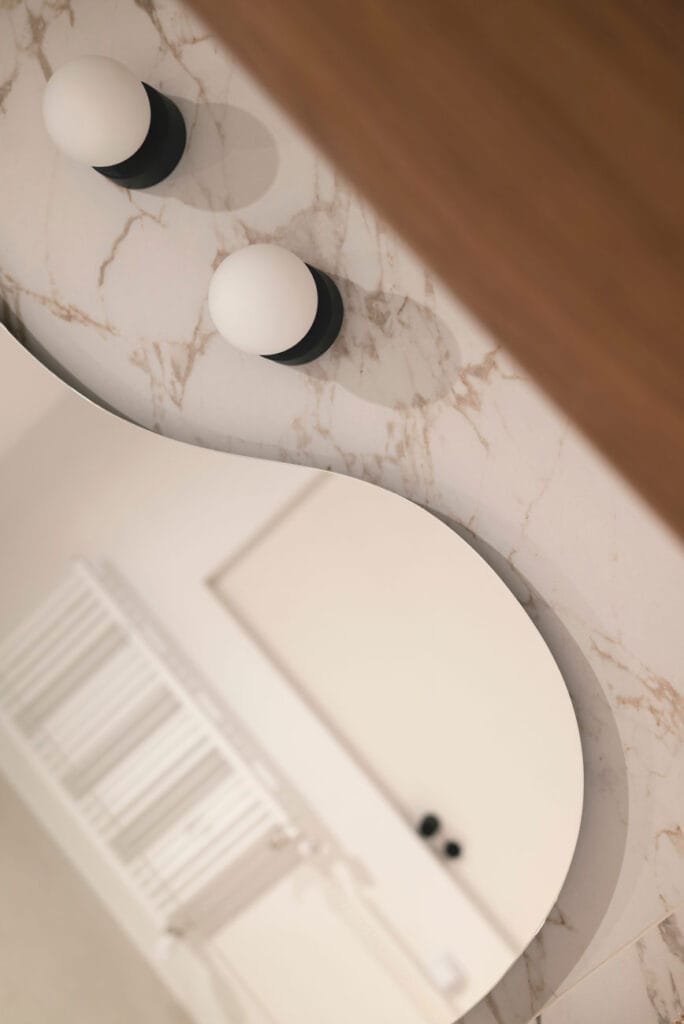 Odbicie wnętrza łazienki w nieregularnym lustrze na tle marmurowych ścian, z designerskimi lampami ściennymi.