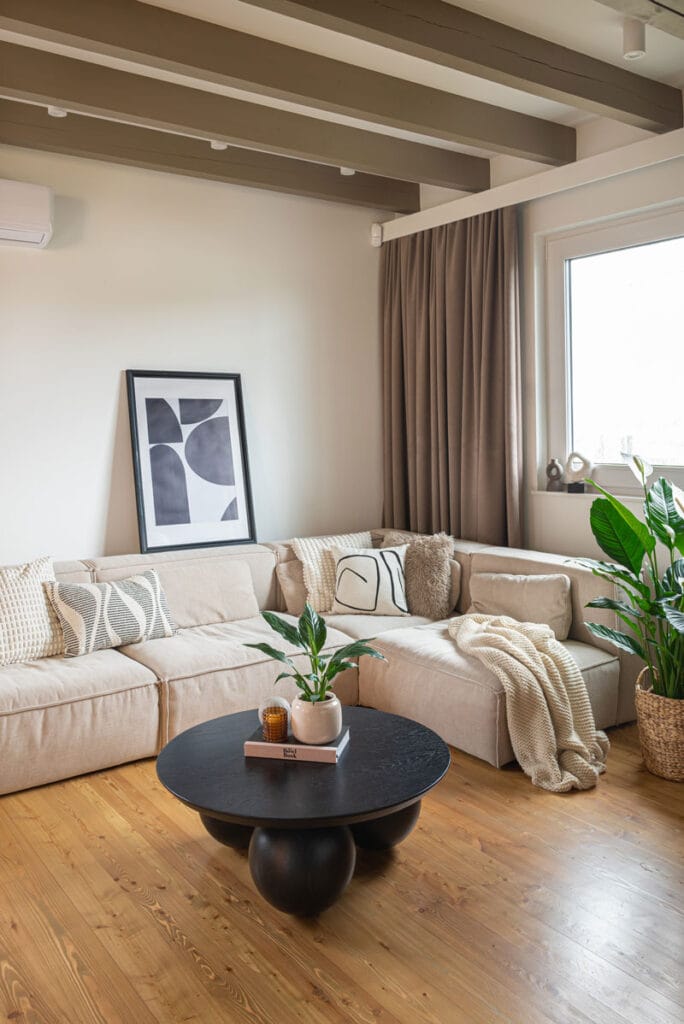 Salon z dużą beżową sofą, czarnym stolikiem kawowym i roślinami w tle, z drewnianymi belkami na suficie.