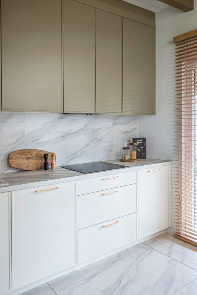 Nowoczesna kuchnia z marmurowym blatem, jasnymi szafkami i subtelnymi złotymi detalami, oświetlona naturalnym światłem.