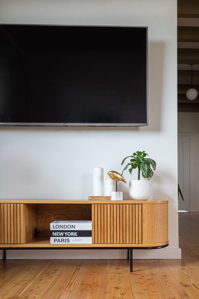 Minimalistyczna aranżacja salonu z telewizorem na ścianie i stylową drewnianą komodą. Na blacie dekoracje: świeczki, złota figurka ptaka i doniczka z zieloną rośliną.