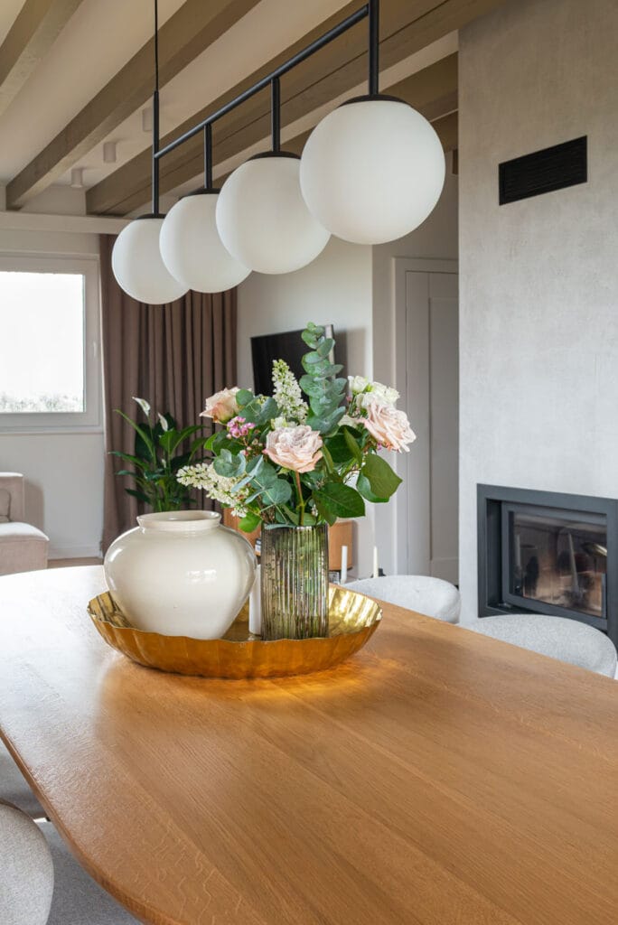 Zbliżenie na dekorację stołu w jadalni – elegancka kompozycja z białej ceramicznej wazy, złotej tacy i bukietu kwiatów. W tle nowoczesna lampa wisząca oraz fragment salonu z telewizorem.